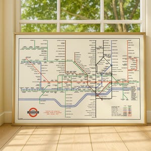 Op de afbeelding: Een ingelijste vintage kaart van de Londense metro, met gekleurde lijnen die metrolijnen voorstellen, met stationsnamen en een rond logo. De kaart zit in een gouden lijst en wordt voor een raam getoond.