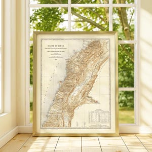 May include: Framed vintage map of Lebanon, featuring detailed geographical information in sepia tones. The map is encased in a gold-colored frame and displayed against a window with a view of greenery. The text "CARTE DU LIBAN" is visible at the top.