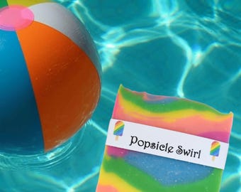 May include: A colourful, rainbow-swirled soap bar with a white label that reads "Popsicle Swirl". The soap is floating in a swimming pool with a beach ball.