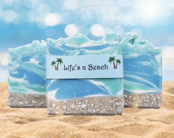 May include: Three handmade soaps with a beach theme. The soaps have blue and white swirls, resembling ocean waves, and a sandy base. A light blue band with the text "Life's a Beach" and palm trees wraps around the center soap. The soaps are on a sandy surface.
