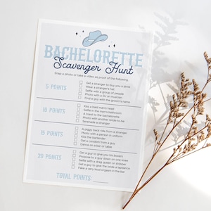 Puede incluir: Una tarjeta de juego de Bachelorette Scavenger Hunt en azul claro y blanco con una ilustración de sombrero de vaquero. La tarjeta enumera tareas con valores de puntos. La tarjeta está sobre una superficie blanca.