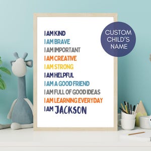 May include: A framed print with the text "I AM KIND, I AM BRAVE, I AM IMPORTANT, I AM CREATIVE, I AM STRONG, I AM HELPFUL, I AM A GOOD FRIEND, I AM FULL OF GOOD IDEAS, I AM LEARNING EVERYDAY, I AM JACKSON". A navy blue circle with the text "CUSTOM CHILD'S NAME" is on the right.