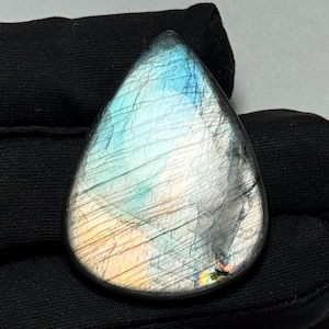 May include: A teardrop-shaped labradorite gemstone featuring a polished surface with visible internal fractures. The stone displays iridescent flashes of blue, gold, and silver light across its greyish base, creating a reflective play of color.