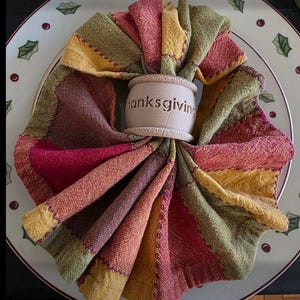 May include: A folded, colorful Thanksgiving napkin arranged on a plate. The napkin is held together by a beige napkin ring with the word "thanksgiving" engraved on it. The napkin features a patchwork design in shades of red, green, and yellow.