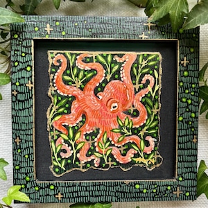 May include: A framed painting of an orange octopus with gold accents surrounded by green foliage on a black background. The frame is green with gold accents and a black background.