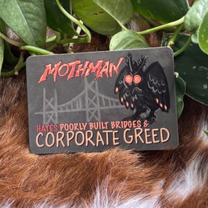 May include: A rectangular Mothman sticker with a gray background. The sticker features a black owl-like creature with red eyes, a bridge illustration, and the text "Mothman Hates Poorly Built Bridges & Corporate Greed."