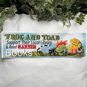 May include: A colourful sticker with the text "Frog and Toad Support Their Local Library & Read Banned Books". The sticker features an illustration of two frogs dressed in punk rock clothing. One frog is reading a book while the other is lying down.