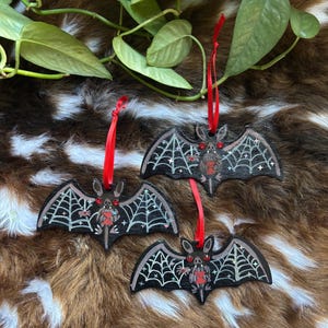 Op de afbeelding: Drie zwarte vleermuisornamenten met witte spinnenwebben en rode accenten. Elke vleermuis heeft een rood lint om op te hangen. De vleermuizen zijn versierd met kleine rode edelstenen en een rood hartontwerp. De ornamenten worden getoond op een bruin en wit patroon.