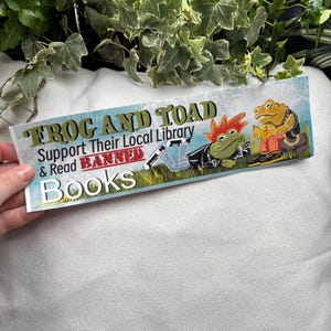 May include: A sticker featuring a green frog and a brown toad reading books. The sticker reads "Frog and Toad Support Their Local Library & Read Banned Books".