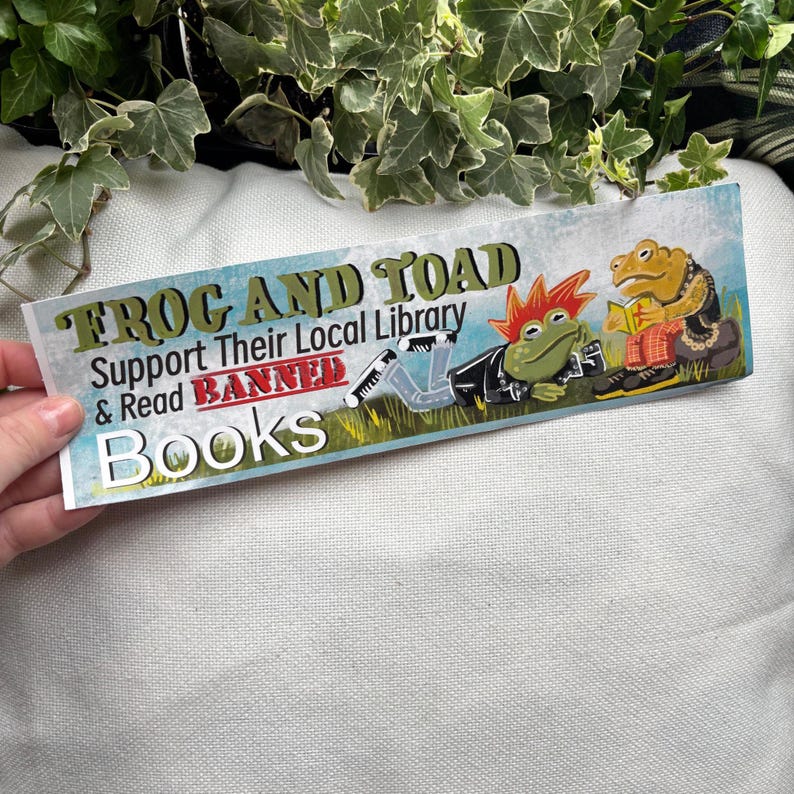 May include: A sticker featuring a green frog and a brown toad reading books. The sticker reads "Frog and Toad Support Their Local Library & Read Banned Books".