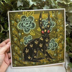 Pu&ograve; includere: Stampa artistica quadrata con un coniglio nero con accenti floreali gialli e occhi rossi. Il coniglio &egrave; ambientato su uno sfondo verde scuro con fiori e foglie stilizzati. La stampa ha uno stile illustrativo stravagante.