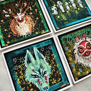 Puede incluir: Cuatro impresiones cuadradas con ilustraciones de personajes de la pel&iacute;cula animada 'La Princesa Mononoke'. Las impresiones est&aacute;n en papel blanco con bordes negros y detalles dorados. Los personajes incluyen un lobo, un esp&iacute;ritu del bosque y un esp&iacute;ritu enmascarado.