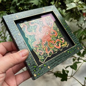 May include: A framed artwork featuring a red octopus with green tentacles surrounded by green foliage on a black background. The frame is green with gold accents and a pattern of green dots and crosses.