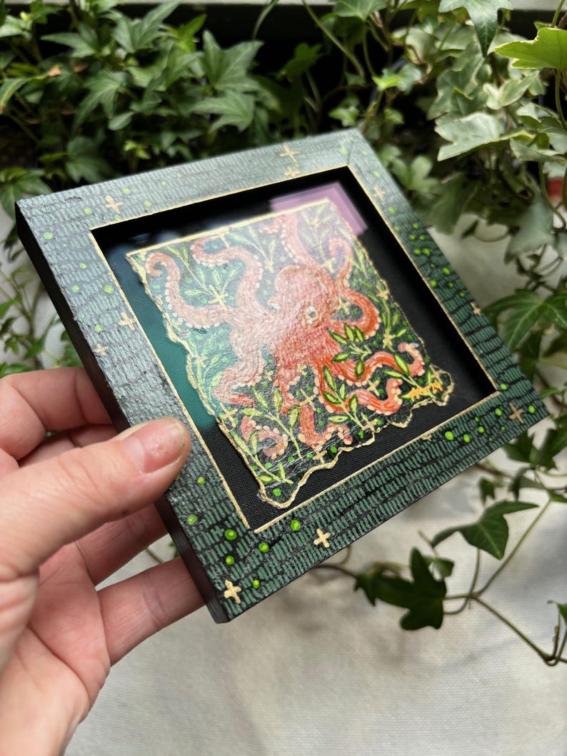 May include: A framed artwork featuring a red octopus with green tentacles surrounded by green foliage on a black background. The frame is green with gold accents and a pattern of green dots and crosses.