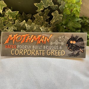 May include: A sticker featuring a cartoon Mothman with glowing eyes, the text "Mothman Hates Poorly Built Bridges & Corporate Greed", and a drawing of a bridge with the text "*?! G!" above it.