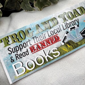 May include: A green and white sticker with the text "Frog and Toad Support Their Local Library & Read Banned Books". The sticker has a red strikethrough over the word "Banned". The sticker also features a cartoon frog wearing a graduation cap and gown.