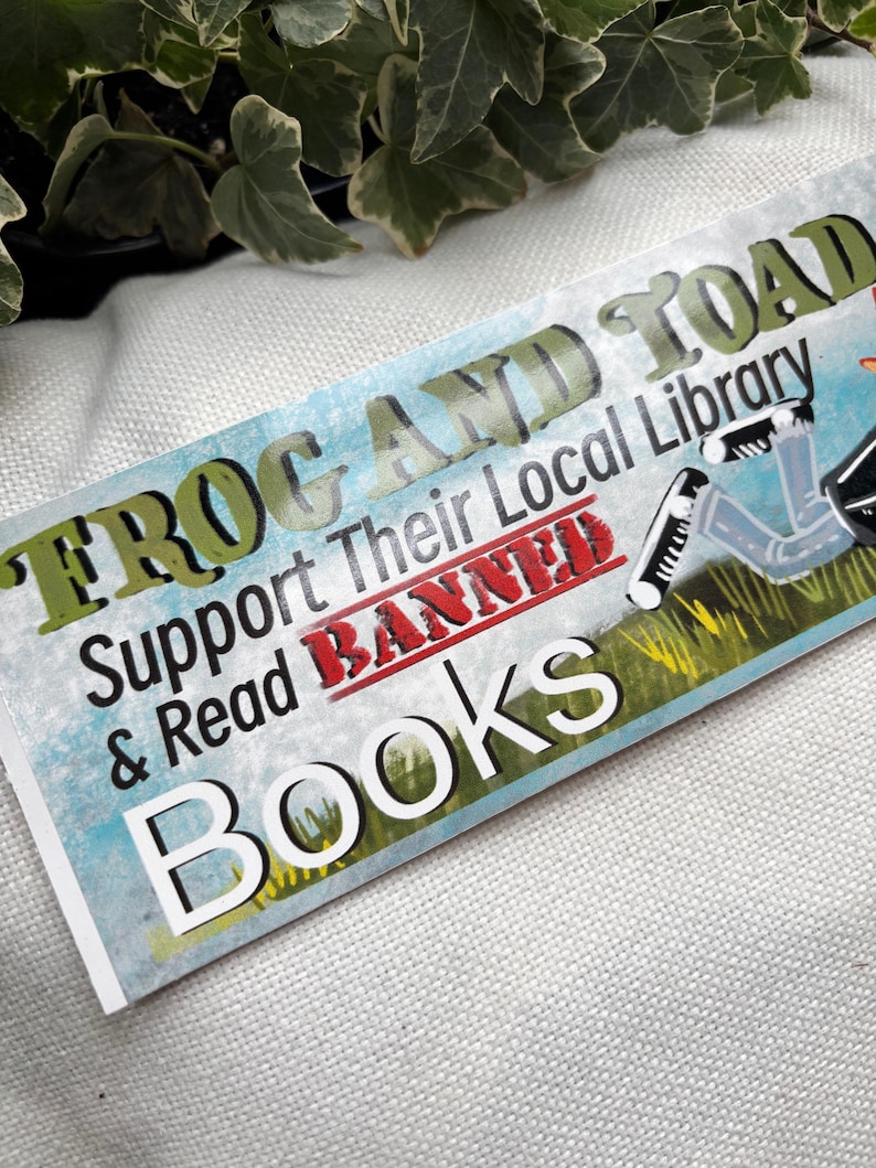 May include: A green and white sticker with the text "Frog and Toad Support Their Local Library & Read Banned Books". The sticker has a red strikethrough over the word "Banned". The sticker also features a cartoon frog wearing a graduation cap and gown.