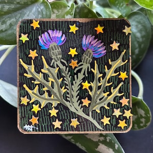 May include: A square, hand-painted wooden art piece featuring two purple thistle flowers with green stems and yellow spiky leaves. The background is black with vertical green lines and yellow star accents. The edges are brown.