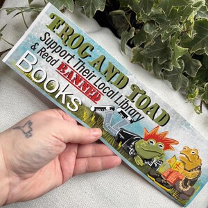 May include: A sticker featuring a green frog and a toad dressed in punk rock attire. The sticker reads "Frog and Toad Support Their Local Library Banned Books".