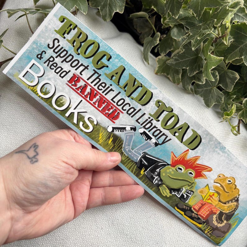 May include: A sticker featuring a green frog and a toad dressed in punk rock attire. The sticker reads "Frog and Toad Support Their Local Library Banned Books".