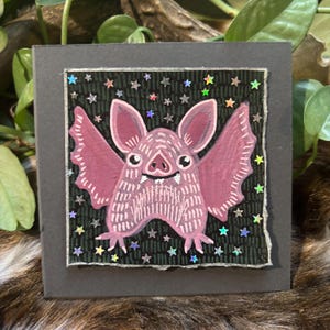 May include: A square, dark gray artwork featuring a pink bat with outstretched wings and a smiling face. The bat is set against a black background with a pattern of small stars. The artwork is framed with a silver border.