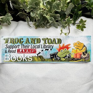May include: A sticker with a blue, green, and yellow background featuring two frogs. The text reads "Frog and Toad Support Their Local Library & Read Banned Books".