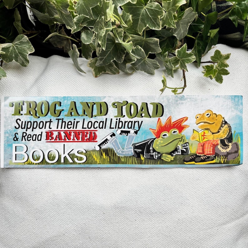 May include: A sticker with a blue, green, and yellow background featuring two frogs. The text reads "Frog and Toad Support Their Local Library & Read Banned Books".