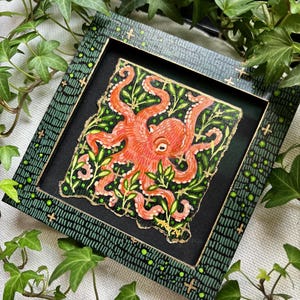 May include: A framed painting of an orange octopus with green leaves on a black background. The frame is green with gold accents and a black background.