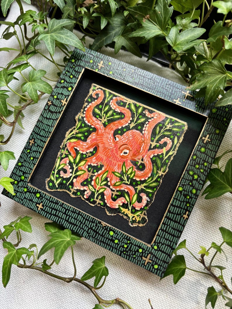 May include: A framed painting of an orange octopus with green leaves on a black background. The frame is green with gold accents and a black background.