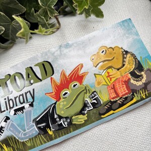 May include: A sticker featuring a green, red, and yellow frog wearing a black leather jacket and blue jeans. The frog is lying down and has a mohawk. The text "Toad Library" is above the frog. A second frog is sitting up and wearing a red and black plaid shirt and a gold chain. The frog is holding a book.
