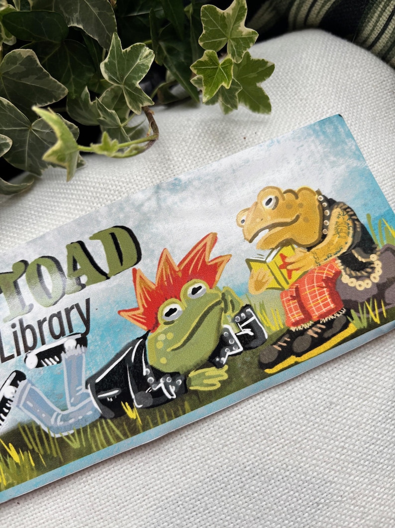 May include: A sticker featuring a green, red, and yellow frog wearing a black leather jacket and blue jeans. The frog is lying down and has a mohawk. The text "Toad Library" is above the frog. A second frog is sitting up and wearing a red and black plaid shirt and a gold chain. The frog is holding a book.