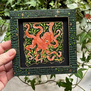 May include: A framed painting of an orange octopus with tentacles surrounded by green leaves on a black background. The frame is green with gold accents.