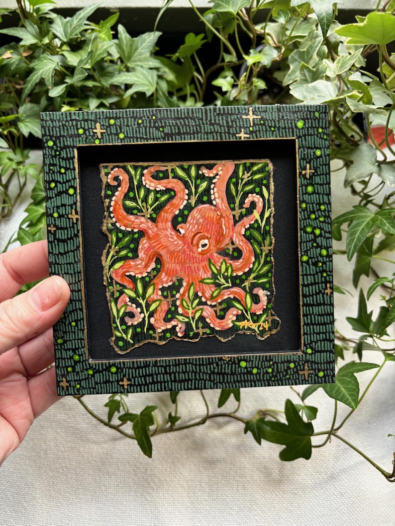 May include: A framed painting of an orange octopus with tentacles surrounded by green leaves on a black background. The frame is green with gold accents.