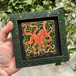 May include: A framed painting of an orange octopus with green foliage on a black background. The frame is green with gold accents.