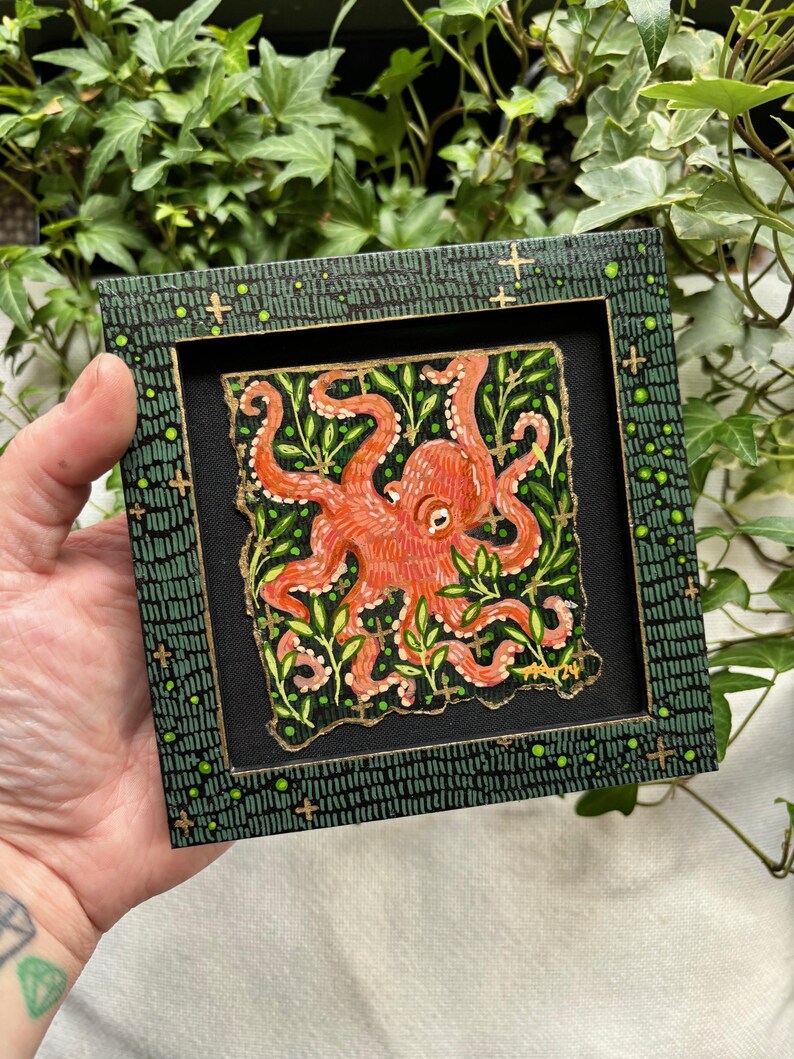 May include: A framed painting of an orange octopus with green foliage on a black background. The frame is green with gold accents.