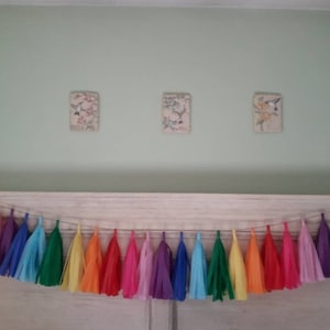 May include: A rainbow-colored tissue paper tassel garland hangs from a white shelf. The garland is made of 20 tassels in various colors, including red, orange, yellow, green, blue, indigo, and violet. The tassels are strung together with a thin white string.