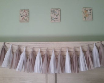 May include: A string of tissue paper tassels in white and beige hangs from a wooden shelf. The tassels are arranged in a repeating pattern of white and beige.