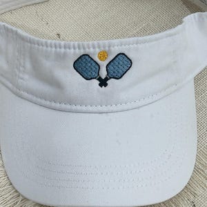 May include: White visor with an embroidered design of two blue pickleball paddles and a yellow ball.