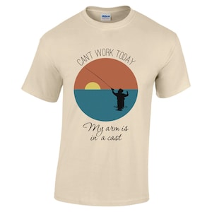 May include: A beige t-shirt featuring a graphic of a person fishing at sunset. The text "Can't work today, my arm is in a cast" is printed on the shirt.