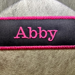 May include: A black fabric patch with a pink embroidered name "Abby" on it. The patch has a pink stitched border.
