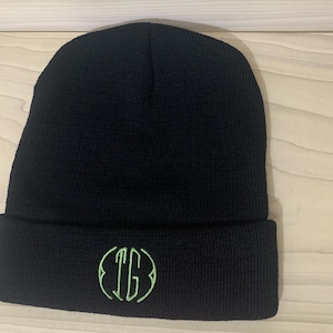 Può includere: Berretto nero con un monogramma ricamato in verde delle lettere "TG" in un cerchio.