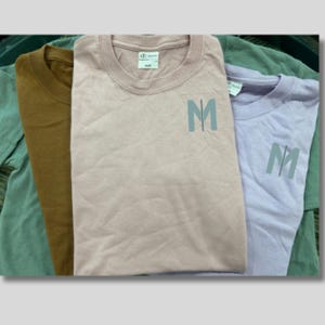 May include: Three crew neck t-shirts in shades of brown, pink, and lavender. Each shirt has a small, light blue letter "M" design on the front. The shirts are folded and stacked, showcasing their colors and design.