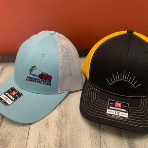 Puede incluir: Dos gorras de camionero negras y amarillas con paneles traseros de malla. Una gorra tiene un logotipo azul y blanco que dice "Wasaga Beach Corvette Club" y la otra gorra tiene un diseño de sol verde.