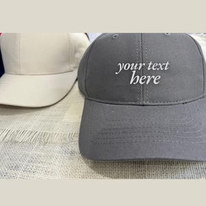 May include: Two baseball caps, one white and one grey. The grey cap has the text "your text here" embroidered on the front.