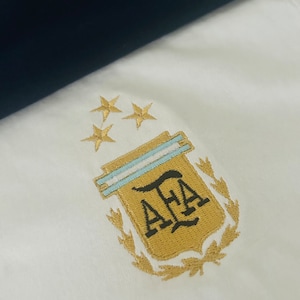 Puede incluir: Tela blanca con un emblema bordado con las letras "AFA" en dorado, una franja azul claro y tres estrellas doradas. En el fondo se ve una tela azul marino oscuro.