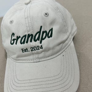 Op de afbeelding: Een beige baseballpet met een groen geborduurd ontwerp met de tekst "Grandpa Est. 2024".