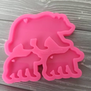 May include: Pink silicone mold with three bear shapes. The largest bear is in the back, with a smaller bear in front of it, and the smallest bear in the front. Each bear has a small heart shape in the center.