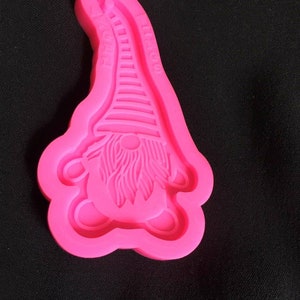 May include: A bright pink silicone mold in the shape of a gnome. The gnome has a long, striped hat, a long beard, and a round nose. The mold is designed for crafting and creating decorative items.