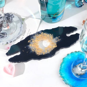 May include: A black and gold resin wine bottle and glass holder with a unique, organic shape. The holder features a central hole for the bottle and two cutouts for wine glasses. Gold flakes are embedded in the resin, adding a touch of elegance.