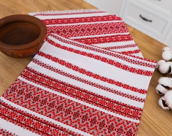 Puede incluir: Un camino de mesa blanco con patrones geométricos bordados en rojo. El camino de mesa tiene un borde con flecos.